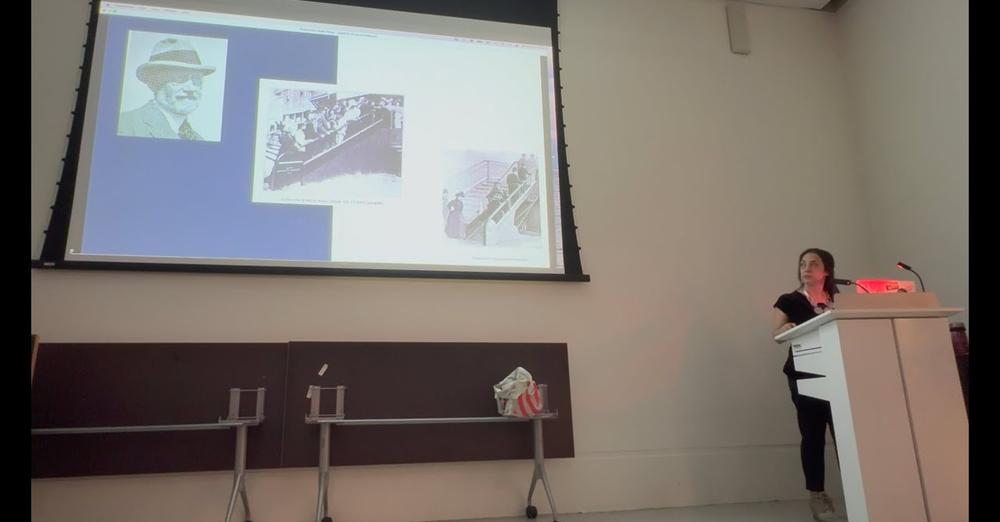 Person at lectern looking up at screen showing escalator information